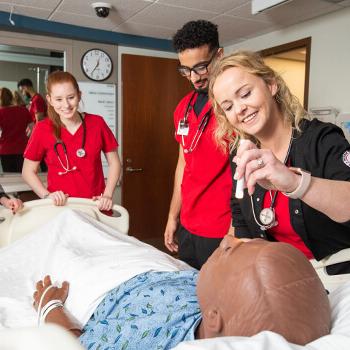 Nursing students work in simulated hospital room; one of them checks the manikin patient’s pupil’s reaction to light.